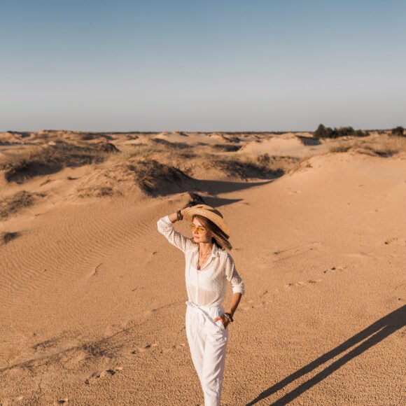 stylish beautiful woman walking in desert sand in white outfit wearing straw hat on sunset, travel safari on vacation